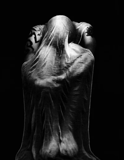 Corps recouvert d’un drap translucide en clair-obscur, posture repliée et chargée d’émotion.
