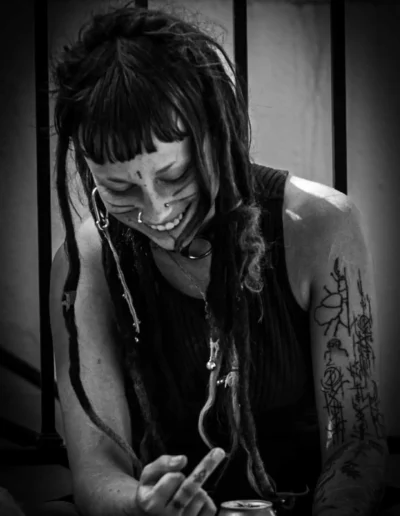 Femme souriante aux dreadlocks et tatouages, photographiée en noir et blanc, exprimant une émotion brute et naturelle.